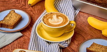 Yellow mug with latte sitting on wood table top with banana bread slices next to it