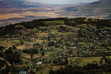 Aerial view of a lush, green agricultural landscape in Kenya's Rift Valley, with scattered homes, farms, and tree-lined plots. In the distance, the terrain flattens into a dry, golden plain bordered by hazy mountain ranges under a muted sky.