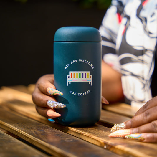 blue tumbler with the words "all are welcome" on it with a rainbow bench